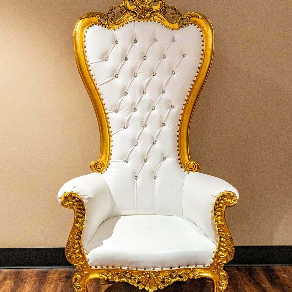 Ornate white and gold throne chair with tufted upholstery and sparkling accents on a wooden floor.