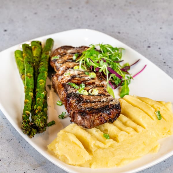Grilled steak with asparagus, mashed potatoes, and a side salad on a white rectangular plate.