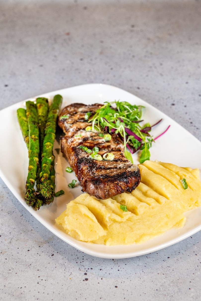 Grilled steak with asparagus, mashed potatoes, and a side salad on a white rectangular plate.