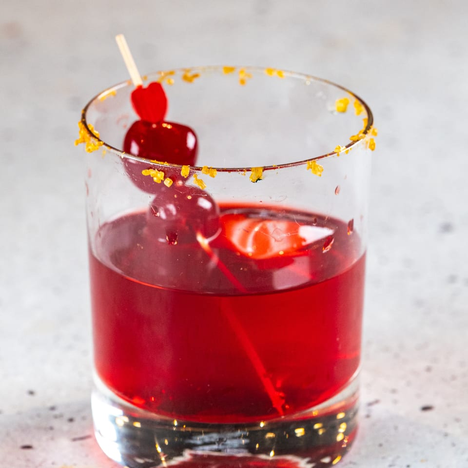 Red cocktail in a glass with an orange sugar rim and cherry garnish on a light background.