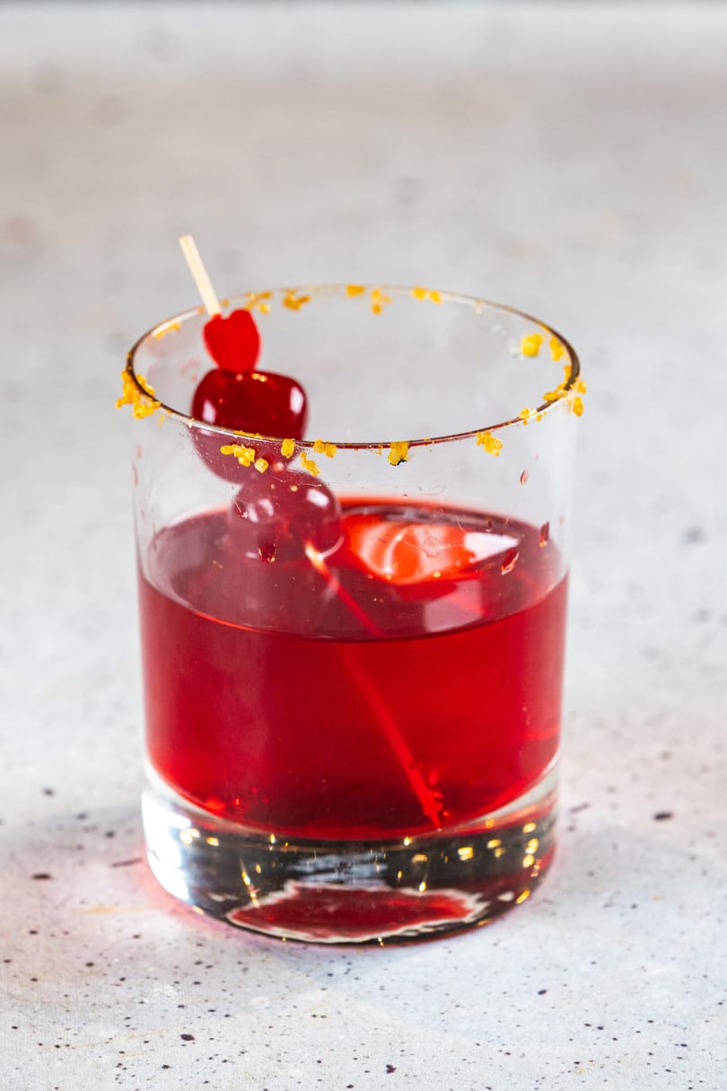Red cocktail in a glass with an orange sugar rim and cherry garnish on a light background.
