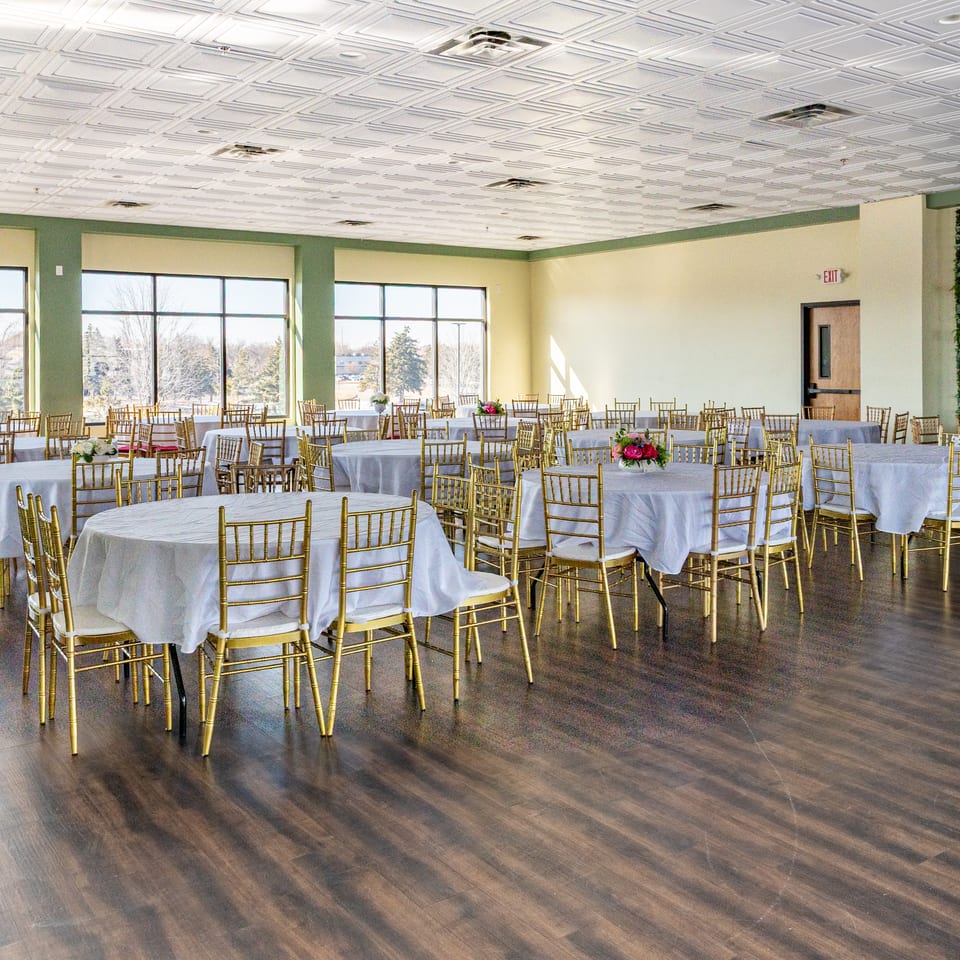 A large, empty banquet hall with round tables, white tablecloths, and gold chairs, ready for an event.