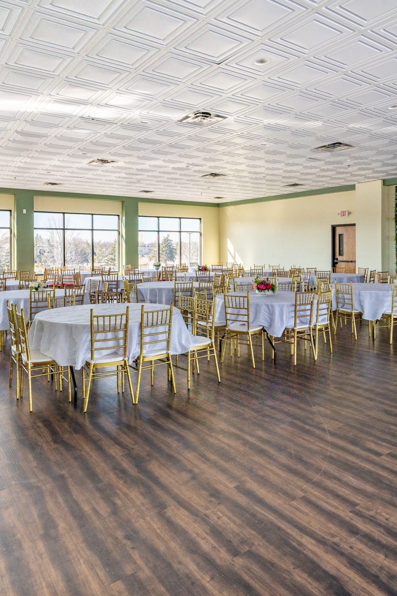 A large, empty banquet hall with round tables, white tablecloths, and gold chairs, ready for an event.