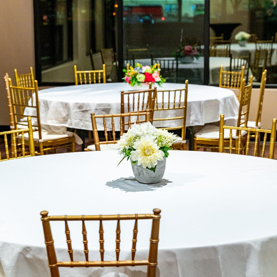 Round tables with white tablecloths and gold chairs, decorated with floral centerpieces, in a banquet hall.