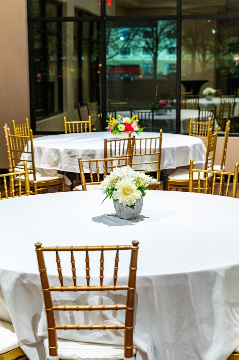 Round tables with white tablecloths and gold chairs, decorated with floral centerpieces, in a banquet hall.