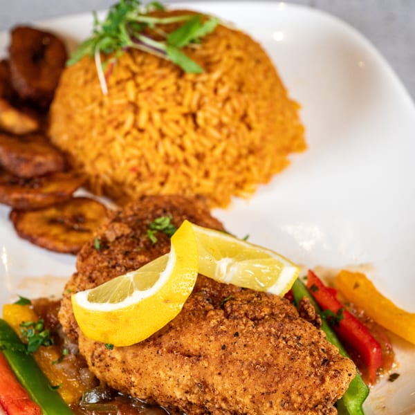 Crispy fried chicken with lemon, jollof rice, plantains, and bell peppers on a white plate.