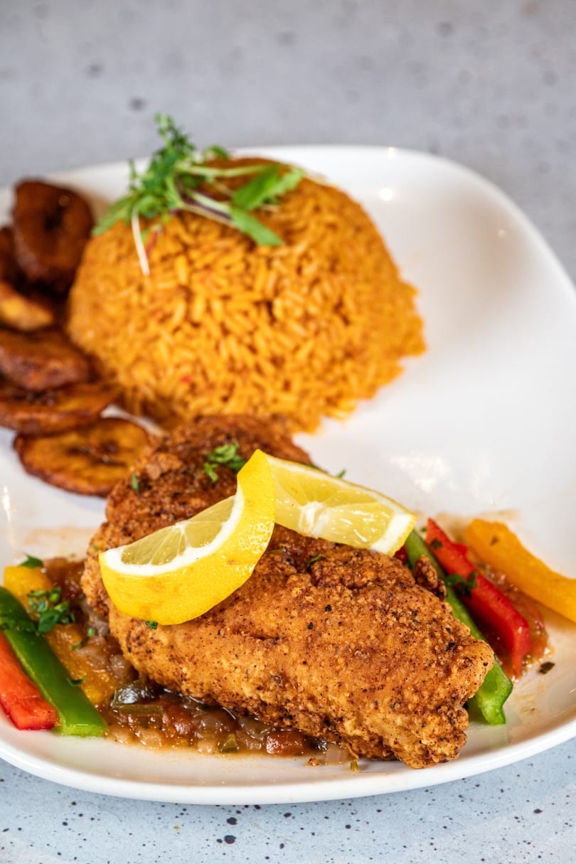 Crispy fried chicken with lemon, jollof rice, plantains, and bell peppers on a white plate.