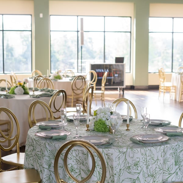 Elegant dining room with round tables set with green and white floral tablecloths and gold chairs.