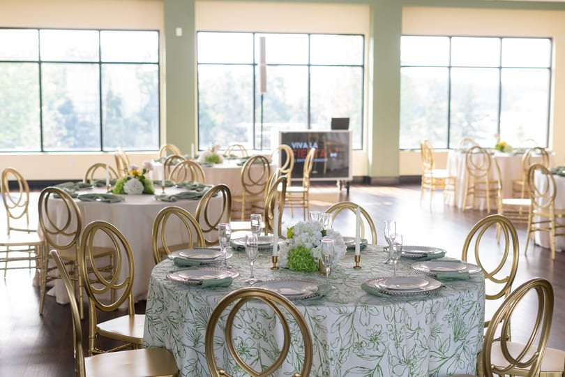 Elegant dining room with round tables set with green and white floral tablecloths and gold chairs.