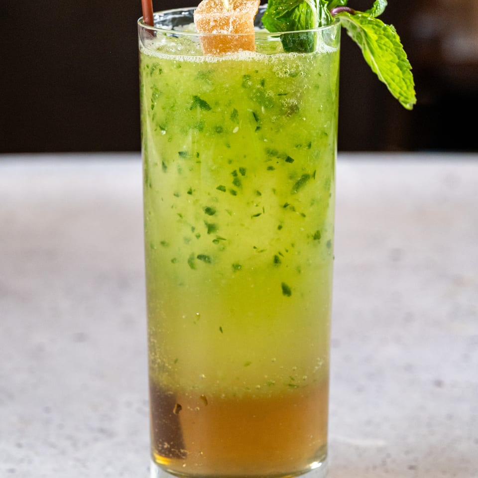 Tall glass of green cocktail with mint leaves, a candied ginger garnish, and a brown straw.