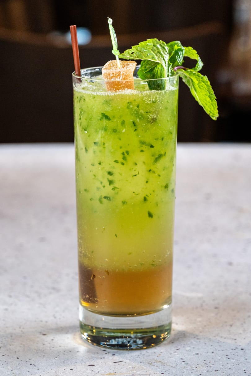 Tall glass of green cocktail with mint leaves, a candied ginger garnish, and a brown straw.