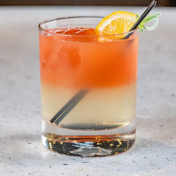A vibrant, layered cocktail with orange and yellow hues, garnished with a lemon slice and black straw.