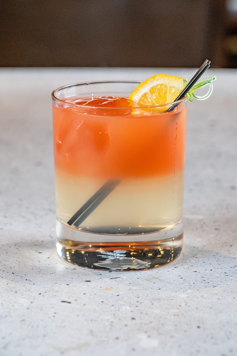 A vibrant, layered cocktail with orange and yellow hues, garnished with a lemon slice and black straw.