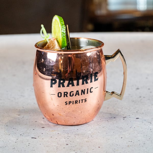 A copper mug with "PRAIRIE ORGANIC SPIRITS" on it, garnished with lime and candied ginger.