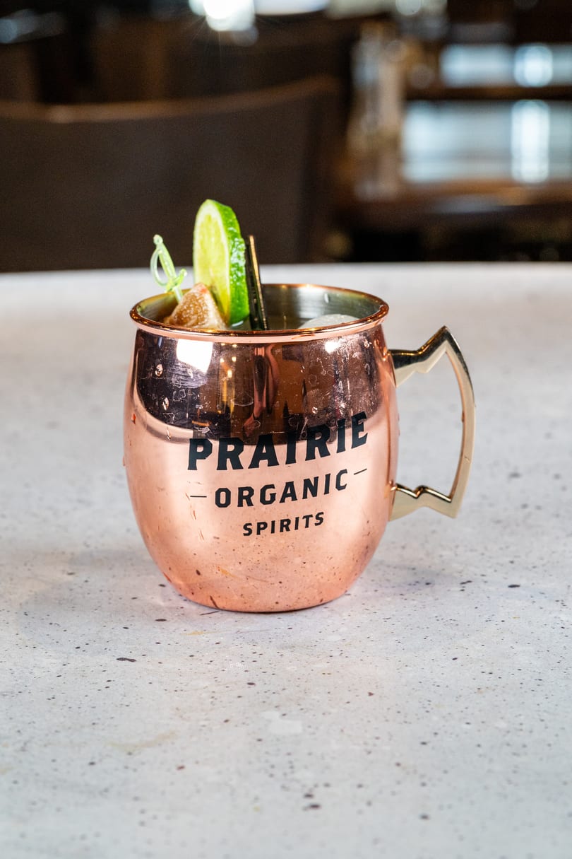 A copper mug with "PRAIRIE ORGANIC SPIRITS" on it, garnished with lime and candied ginger.