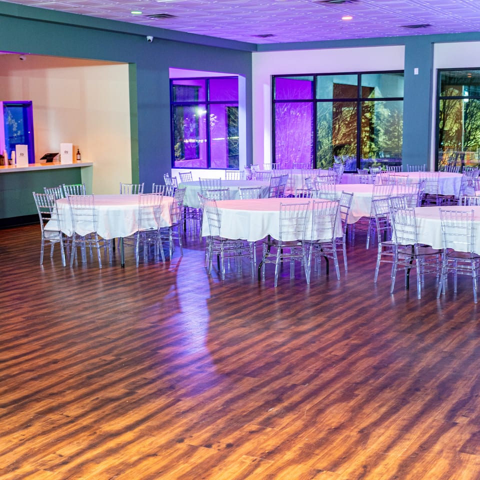 An empty event hall with round tables, clear chairs, and purple lighting on a wooden floor.