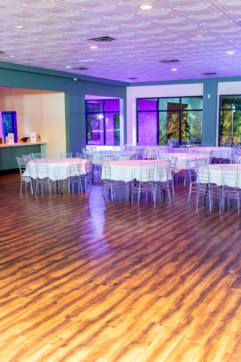 An empty event hall with round tables, clear chairs, and purple lighting on a wooden floor.