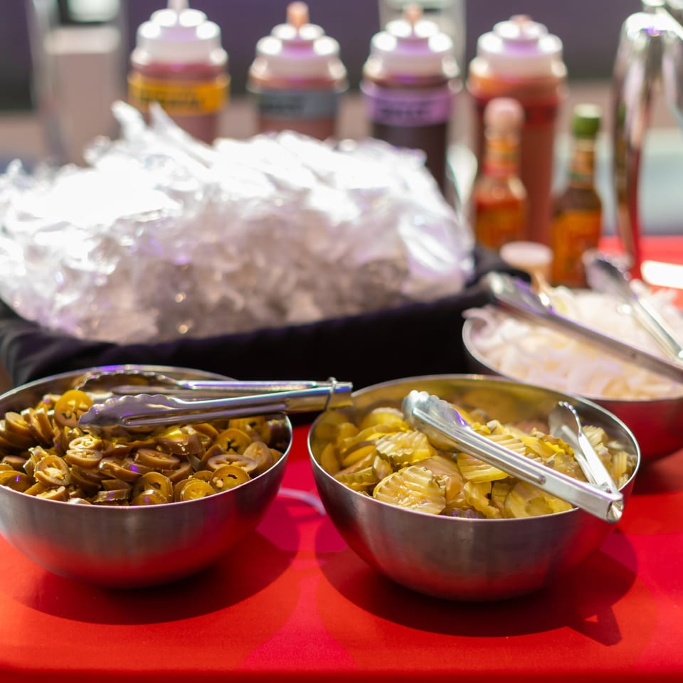 A buffet table with bowls of sliced jalapeños, pickles, onions, and various sauces.