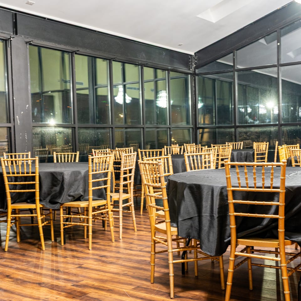 Empty dining room with round tables draped in black, gold chairs, and large windows.