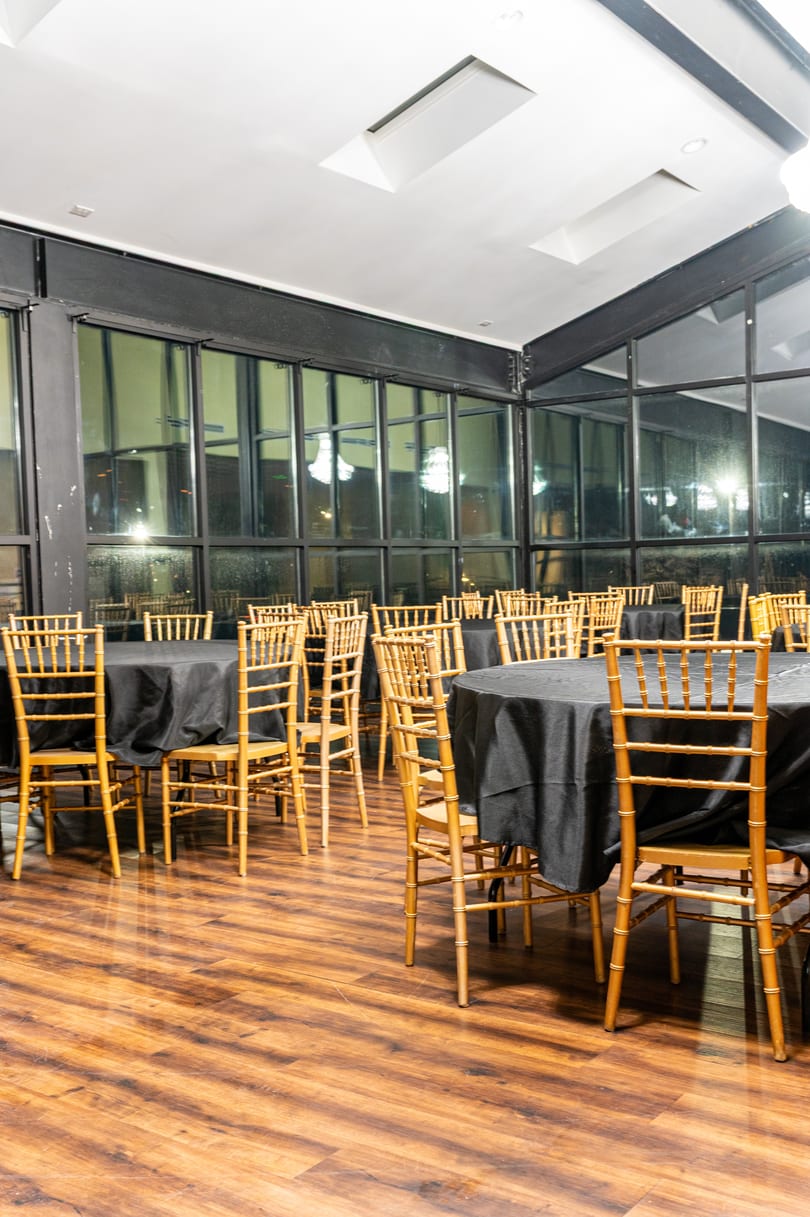 Empty dining room with round tables draped in black, gold chairs, and large windows.