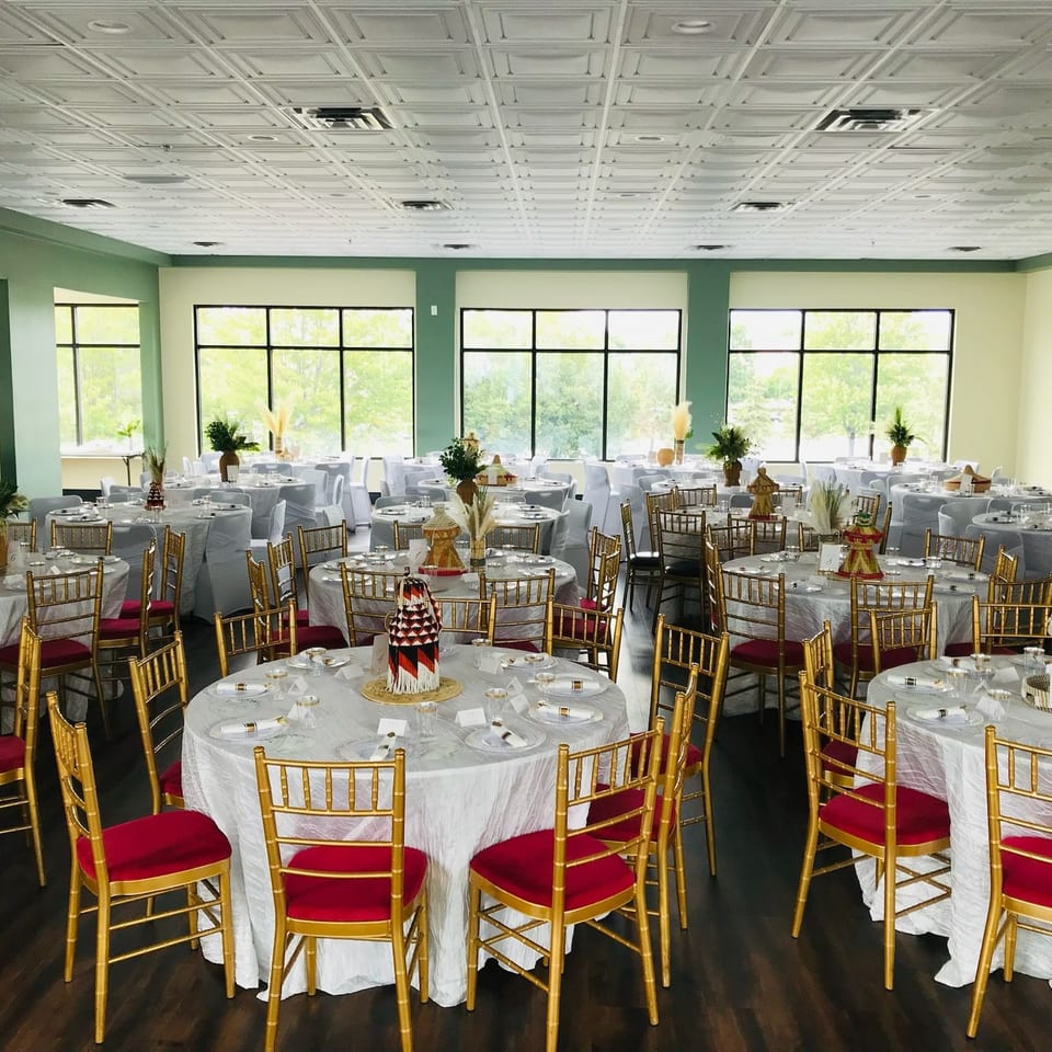 A large dining room with round tables, white tablecloths, gold chairs with red cushions, and large windows.