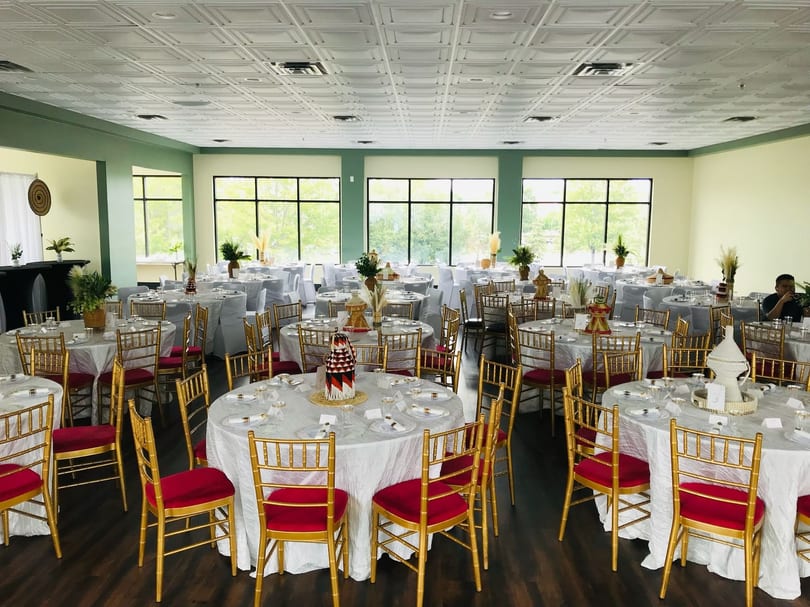 A large dining room with round tables, white tablecloths, gold chairs with red cushions, and large windows.