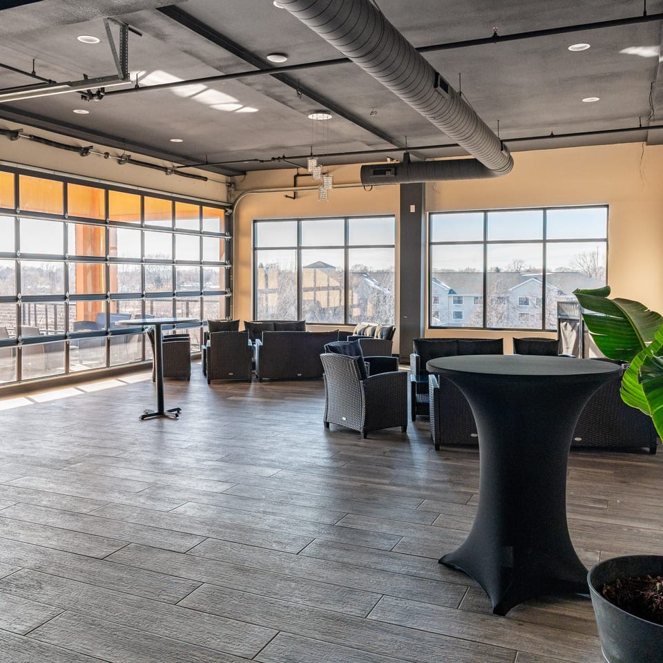 Spacious event room with large windows, modern seating, high-top tables, and exposed ceiling ducts.