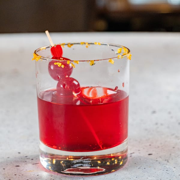 Red cocktail in a glass with a sugared rim and cherry garnish on a light speckled surface.