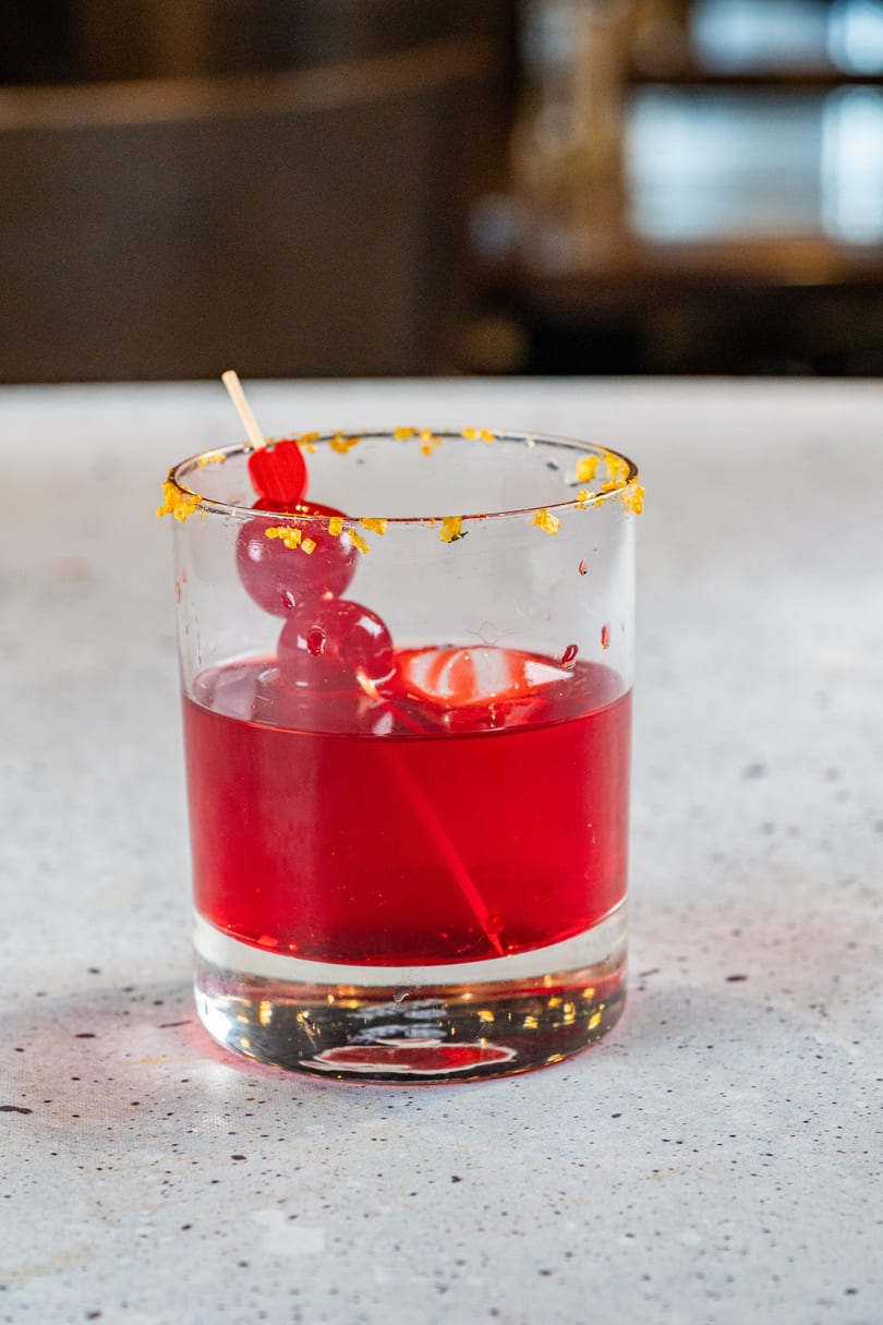 Red cocktail in a glass with a sugared rim and cherry garnish on a light speckled surface.