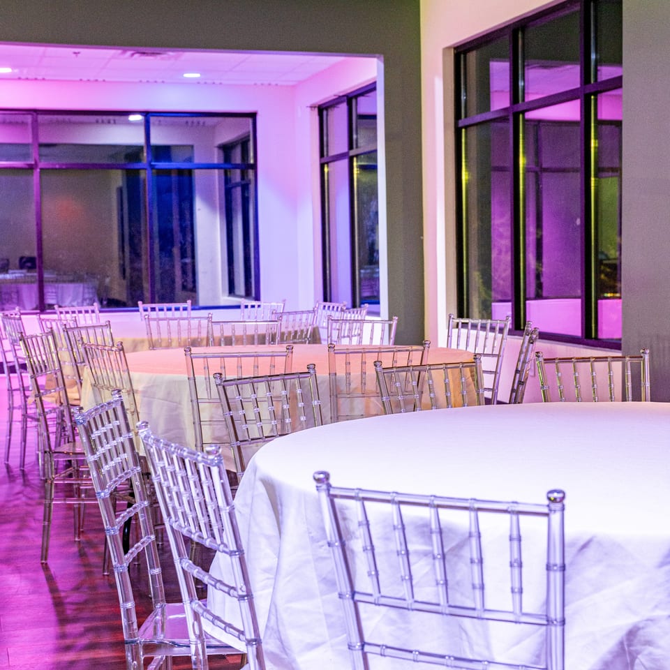 Empty banquet hall with round tables, white tablecloths, and clear chairs under purple lighting.