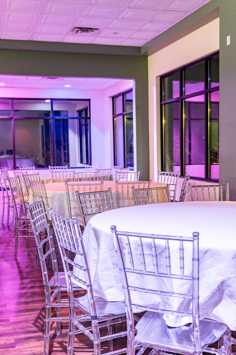 Empty banquet hall with round tables, white tablecloths, and clear chairs under purple lighting.