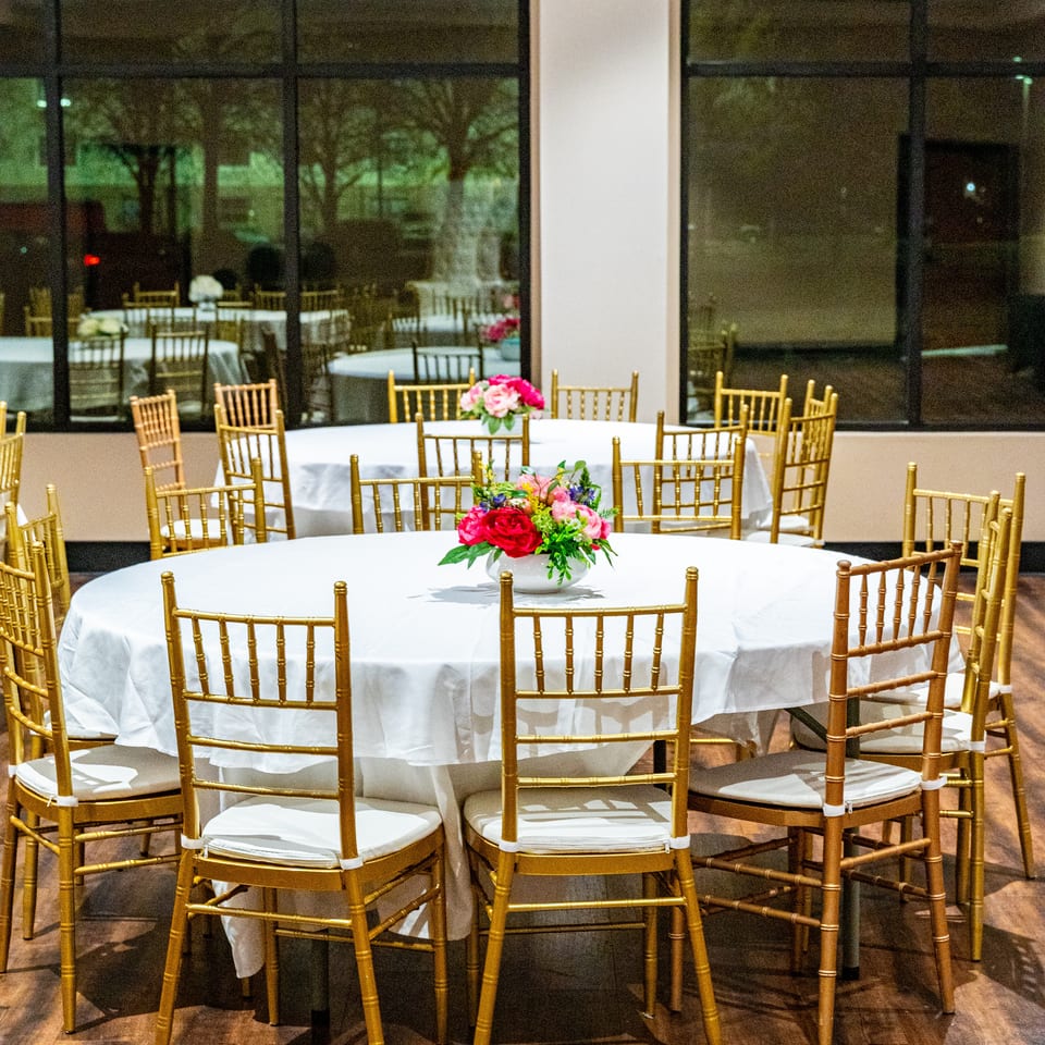 Round tables with white tablecloths and gold chairs, decorated with floral centerpieces, in a banquet hall.