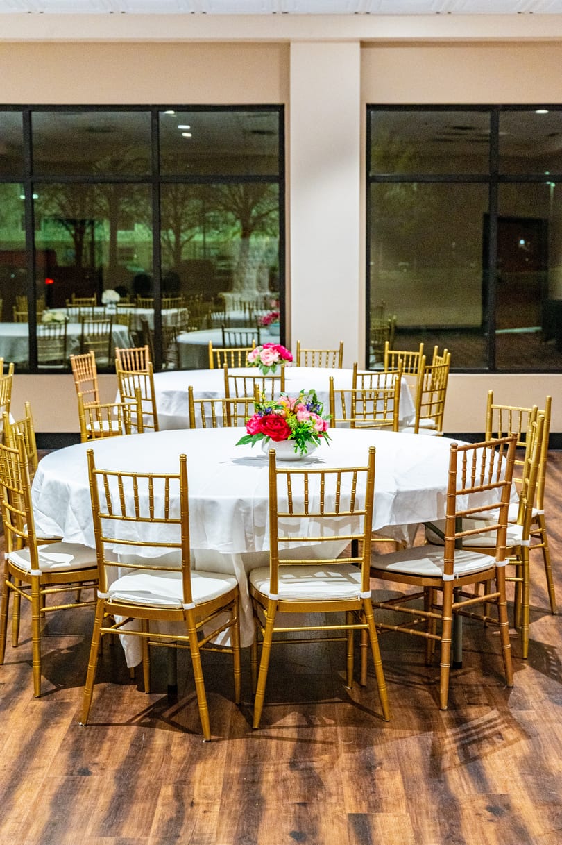Round tables with white tablecloths and gold chairs, decorated with floral centerpieces, in a banquet hall.