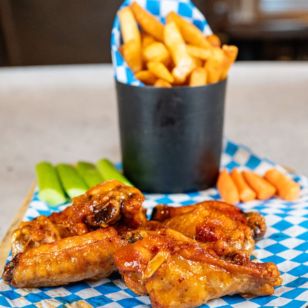 Glazed chicken wings, celery, carrots, and a cup of fries on a blue and white checkered paper.