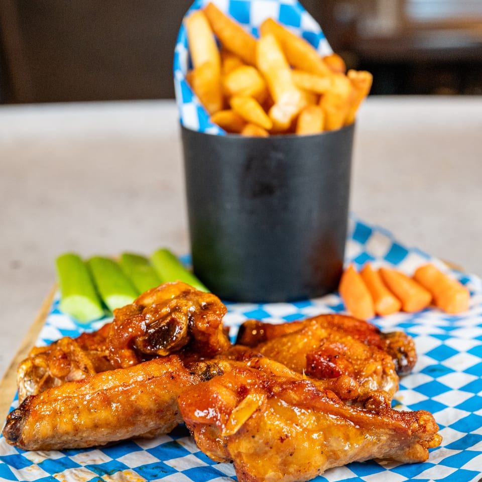 Glazed chicken wings, celery, carrots, and a cup of fries on a blue and white checkered paper.