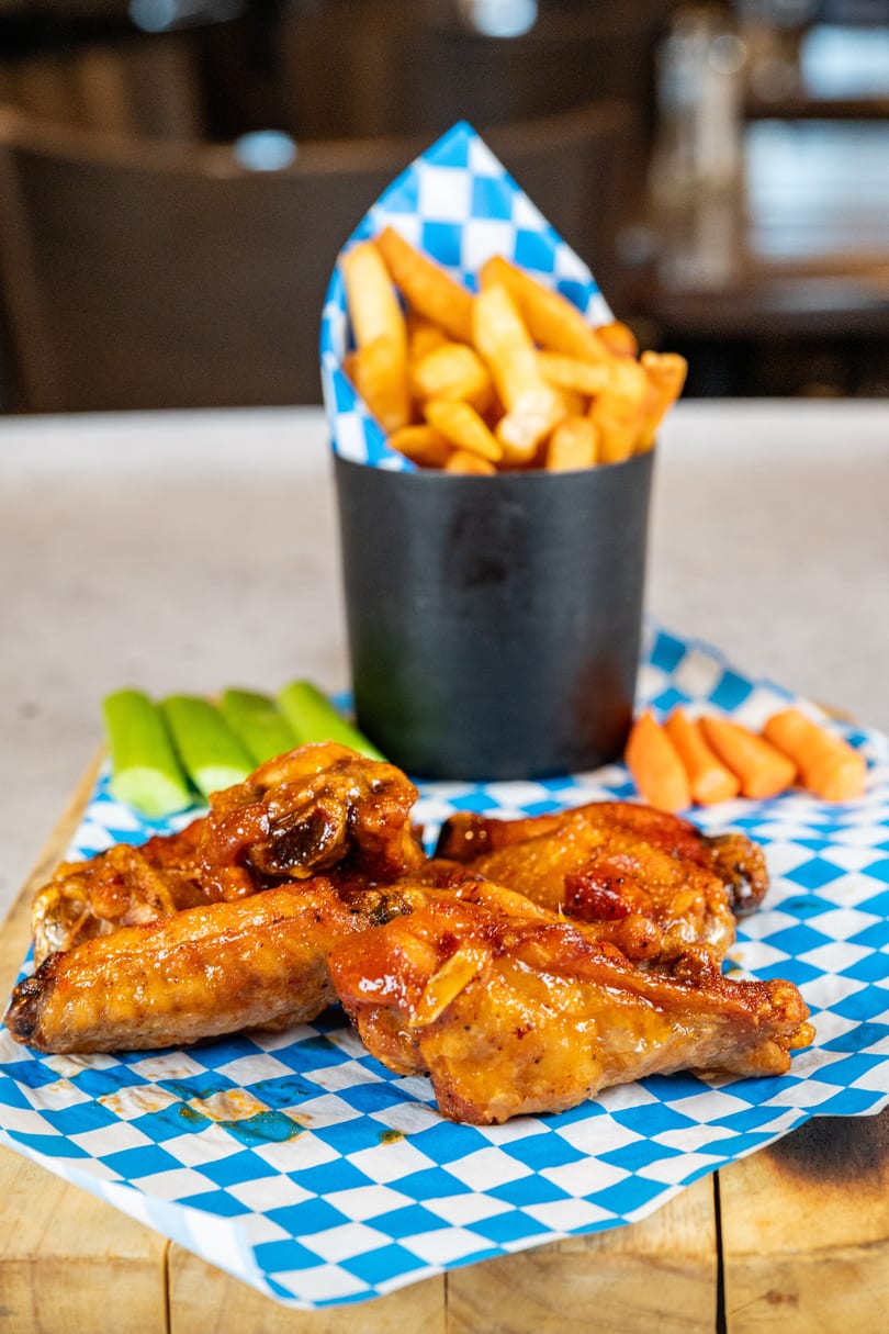 Glazed chicken wings, celery, carrots, and a cup of fries on a blue and white checkered paper.
